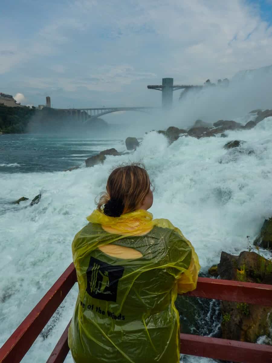 Melissa looking away at the camera wearing a yellow poncho in order to avoid getting wet as she visits Cave of the Winds, in the Niagara Falls beside the Bridal Veil Falls on the New York side. There are rocks everywhere, and the mist from the waterfalls are strong. At a distance, there's a bridge and it's a slightly cloudy day. This is one of the most thrilling and adventurous things to do in Niagara Falls
