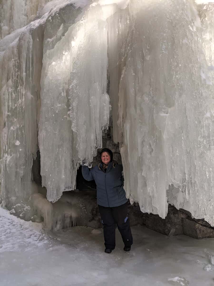 Melissa standing behind a frozen waterfall on the Ice Walk in Maligne Canyon, which look like huge icicles. The ground is covered with snow and ice.