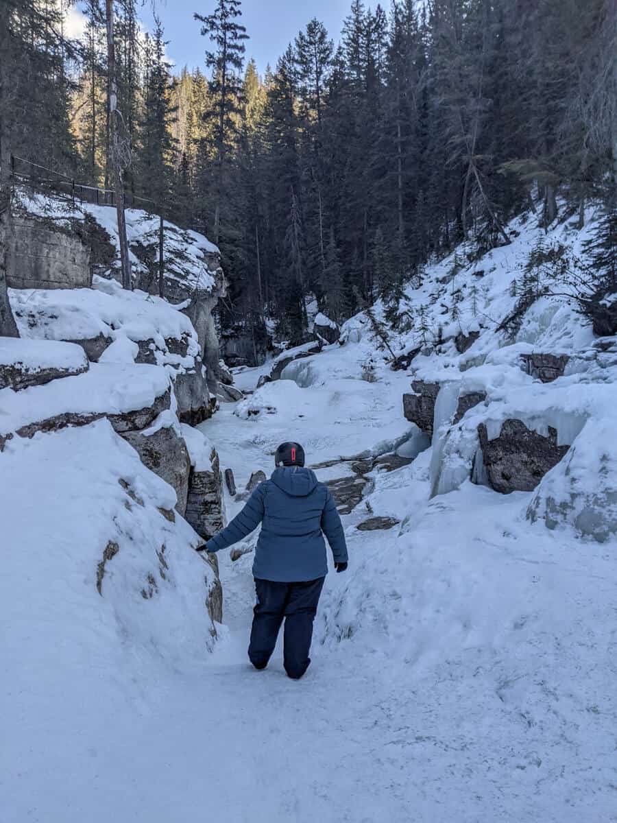 Melissa looking away from the canyon hiking at the third gate in Maligne Canyon Ice Walk located in Jasper National Park, Canada. The ground and rock formations are covered with snow except for a few spots. Beyond the distances, there are some tall trees, with a sunny sky above with minimal clouds
