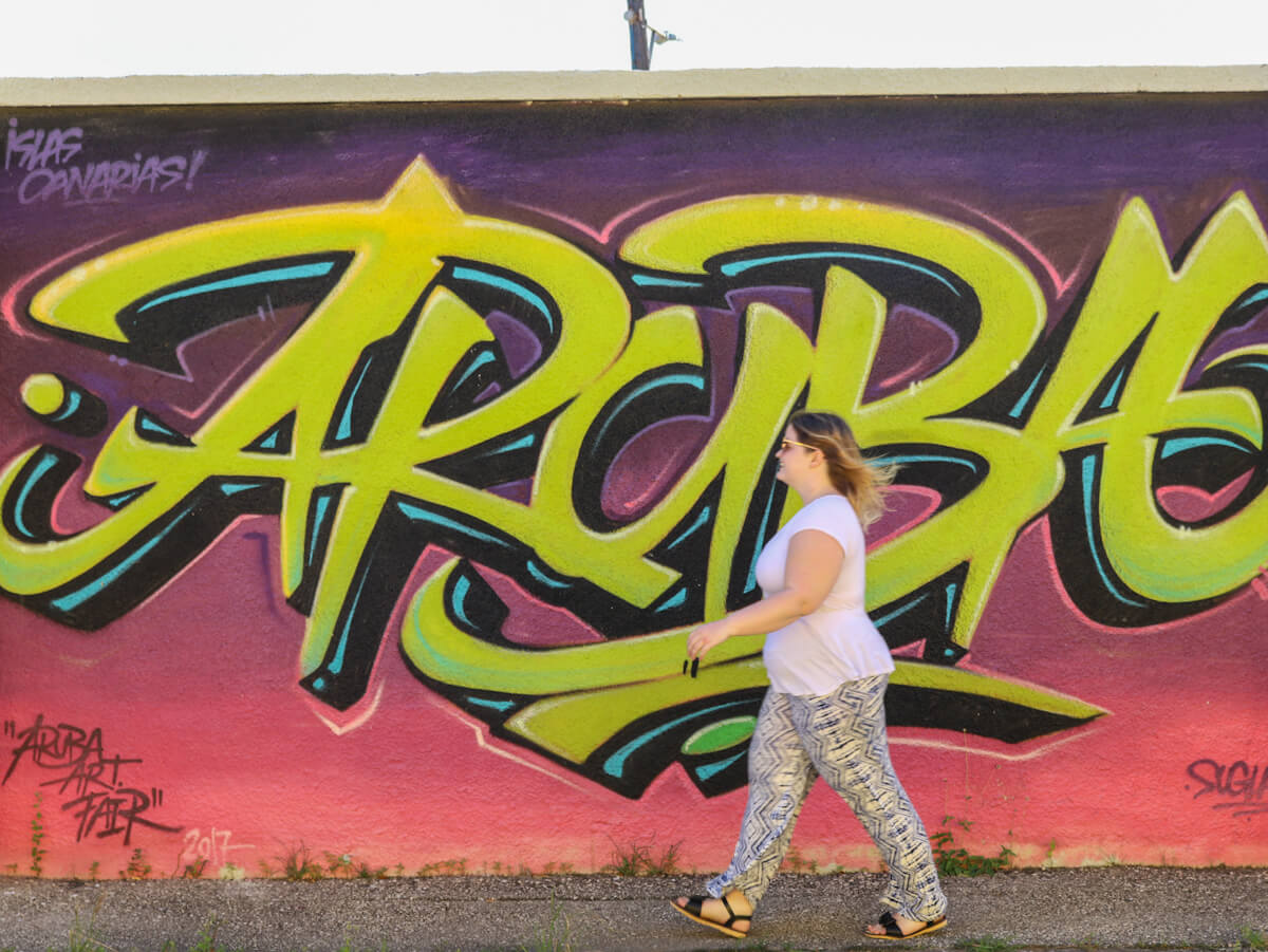 a woman spending a day in Aruba walking in front of a san nicholas street mural that says the phrase aruba in graffiti style writing